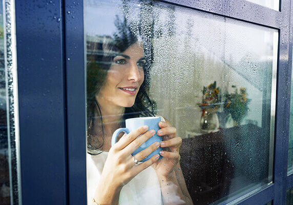 mid adult woman drinking coffee and looking out of the window on rainy day. Horizontal shape